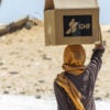 Woman, her head wrapped, balances and carries on her head a box of aid from Gaza Humanitarian Foundation.